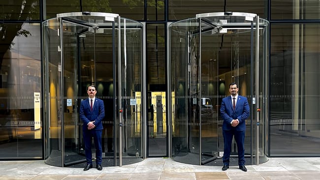 Two male hotel security guards, dressed in Guard Mark uniforms, stand by the glass door, diligently performing their duties.
